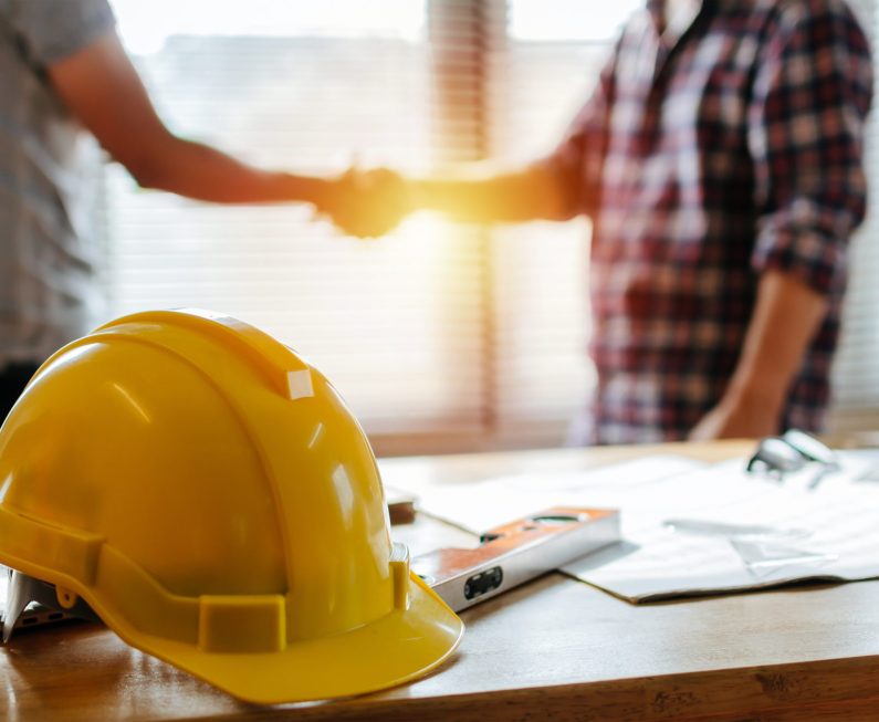 yellow safety helmet on workplace desk with construction worker team hands shaking greeting start up plan new project contract in office center at construction site, partnership and contractor concept